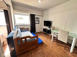 Ein kleines Zimmer mit Bett, Schreibtisch, Stuhl, Fernseher und Fenster.