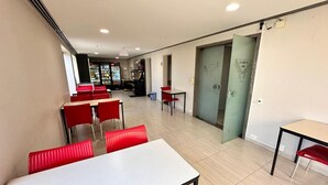 A dining area with tables and chairs, a kitchen in the background, and a door leading to another room. - Le Premier Suites (São Paulo)
