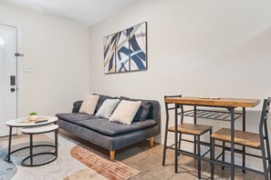 Ein Wohnzimmer mit grauem Sofa, einem kleinen runden Tisch, einem Couchtisch, einem Esstisch mit Stühlen und einem Teppich.