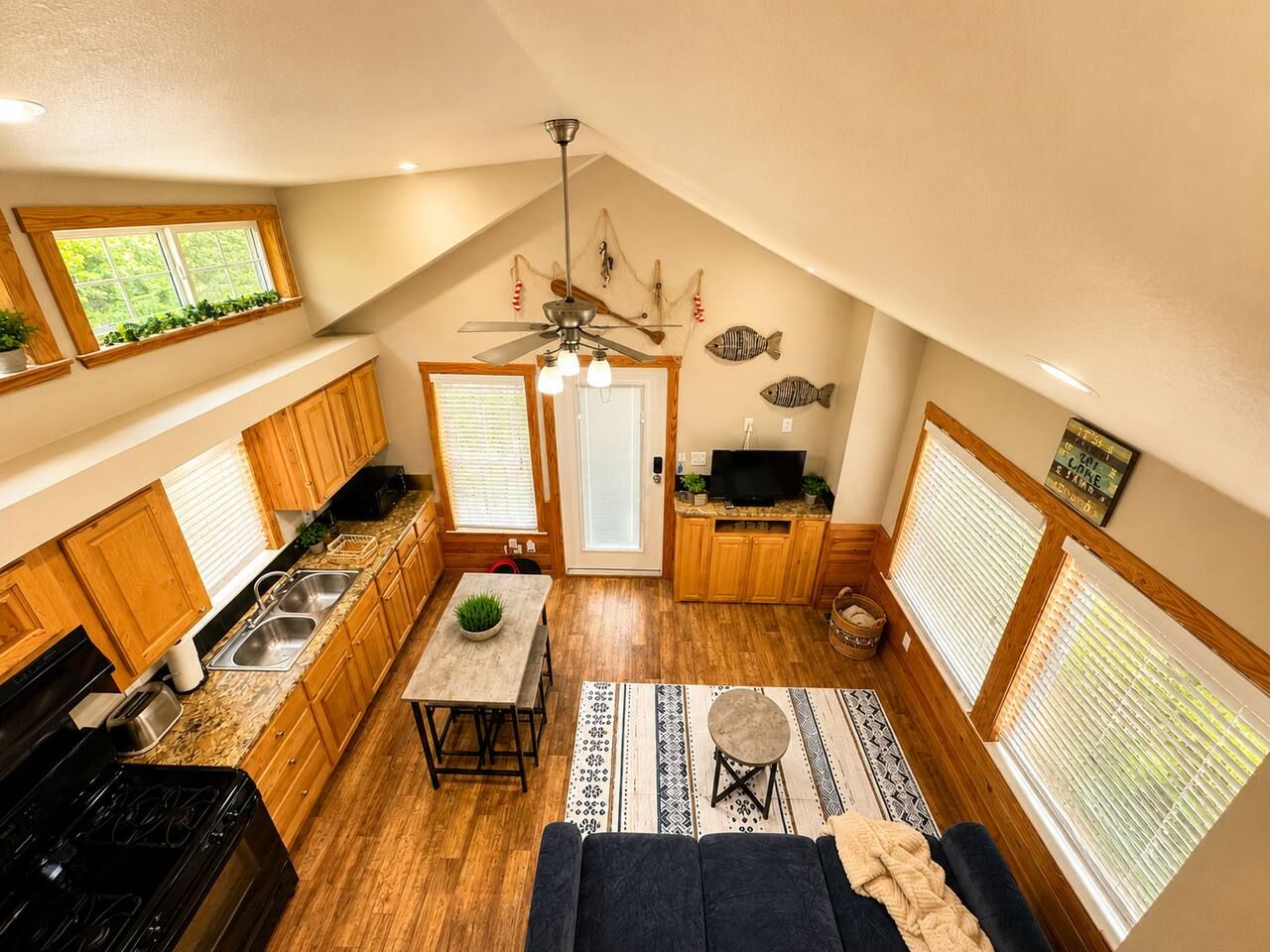 A compact kitchen with wooden cabinets, a dining area with a table and chairs, a ceiling fan, and a television.