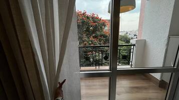 A view through a window with a balcony, a watch on a wrist, and a tree with red flowers.