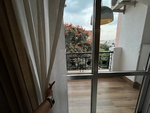 Une vue à travers une fenêtre donnant sur un balcon, une montre au poignet et un arbre aux fleurs rouges.