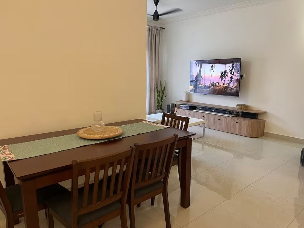 Un coin repas avec une table en bois, des chaises, un téléviseur fixé au mur et, au fond, un espace cuisine.