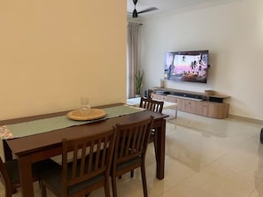 Un coin repas avec une table en bois, des chaises, un téléviseur fixé au mur et, au fond, un espace cuisine.