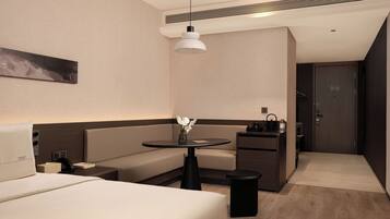 Una habitación de hotel moderna con cama, zona de estar, una mesita con un florero, kitchenette y una puerta.