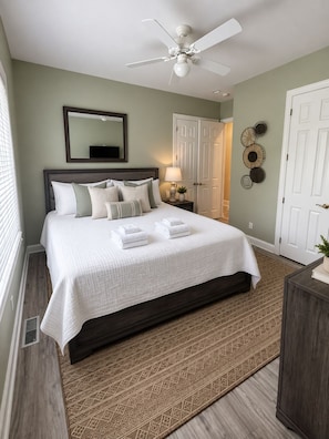 Quarto com cama, ventilador de teto, mesa de cabeceira, espelho e porta.