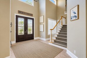 Um corredor com uma porta dupla identificada como 'Beach House', uma escada com corrimão de madeira e um quadro com a imagem de um pássaro na parede.