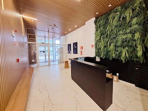 Zona de recepción con techo de madera, un mostrador de recepción y una pared decorada con una instalación de hojas verdes.