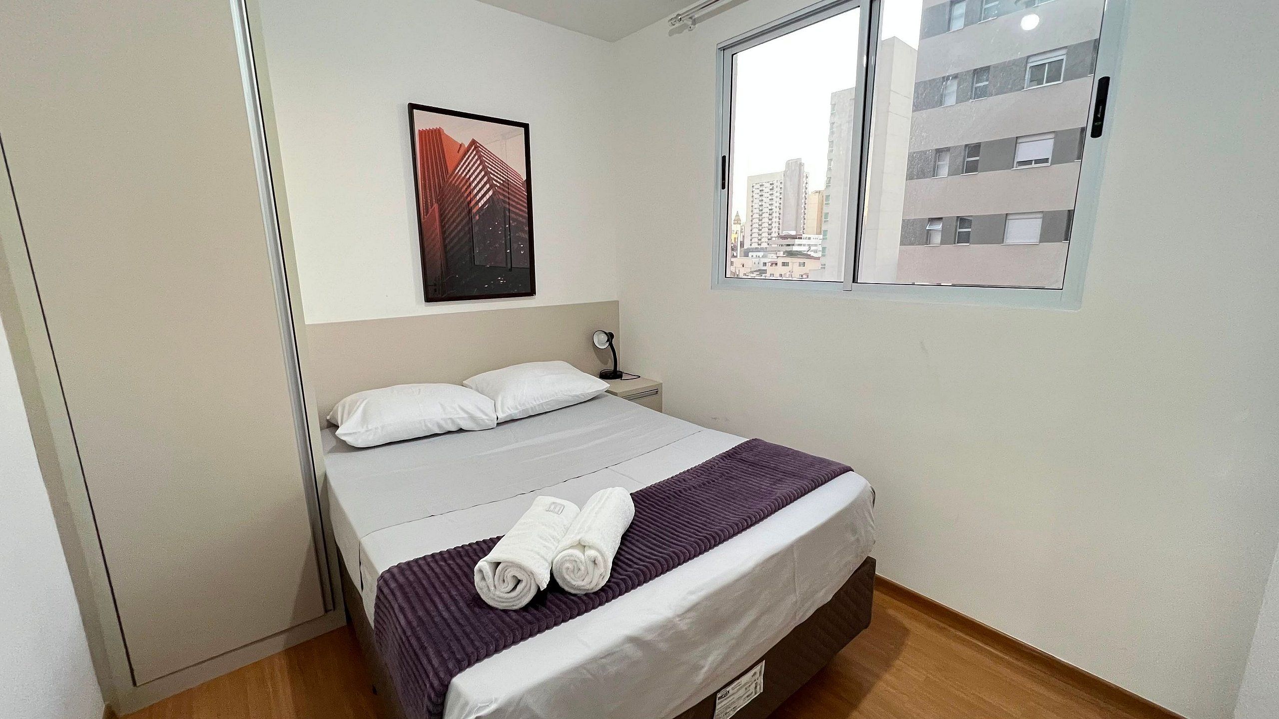 Een hotelkamer met een bed, kussens, een raam met uitzicht op de stad en een lijst met een foto aan de muur.