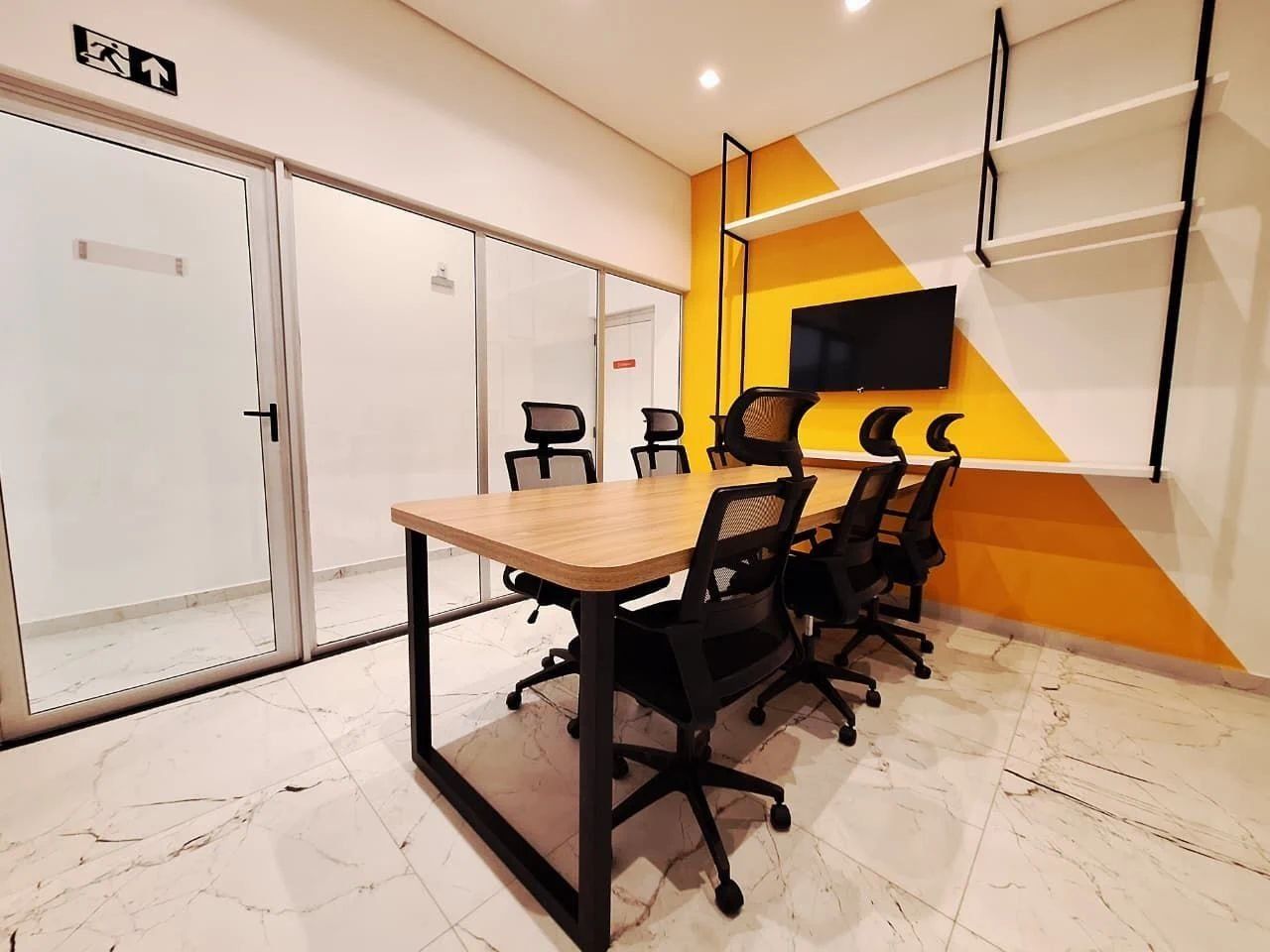 Een vergaderruimte met een lange tafel, zwarte bureaustoelen, een aan de muur bevestigde televisie en een accentmuur in geel.