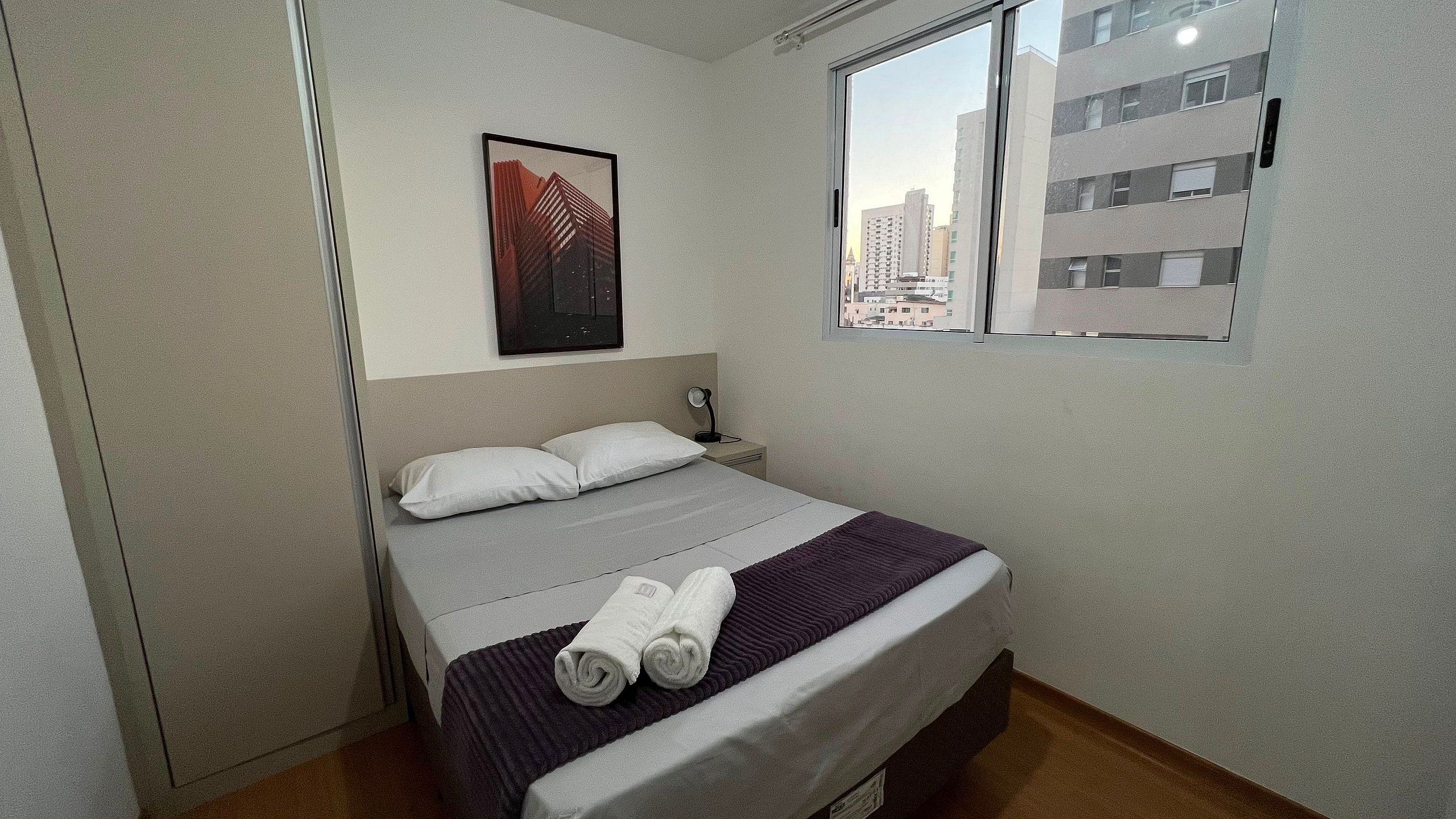 Un lit simple avec une tête de lit, une table de chevet, une lampe et une fenêtre donnant sur des immeubles.