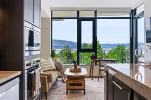 A modern kitchen with a large window, a sofa, and a dining area with a view of the mountains. - The Oak Haven | 2BR+2BA | Central | Pool&Sauna (Kelowna)