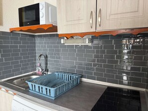 A kitchen with a black tiled backsplash, a stainless steel sink, a dish rack, and wooden cabinets. - "V22" by Versant Collection (Val Thorens)