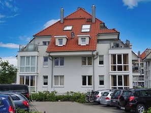 A multi-story residential building with a red-tiled roof, multiple windows, and a parking area with cars and bicycles. - Seestern 32, a family-friendly vacation rental in Großenbrode. An inviting patio with comfortable patio furniture. Wellness oasis with a sauna and hot tub. Storage space for bicycles & seasonal beach chair included (Großenbrode)