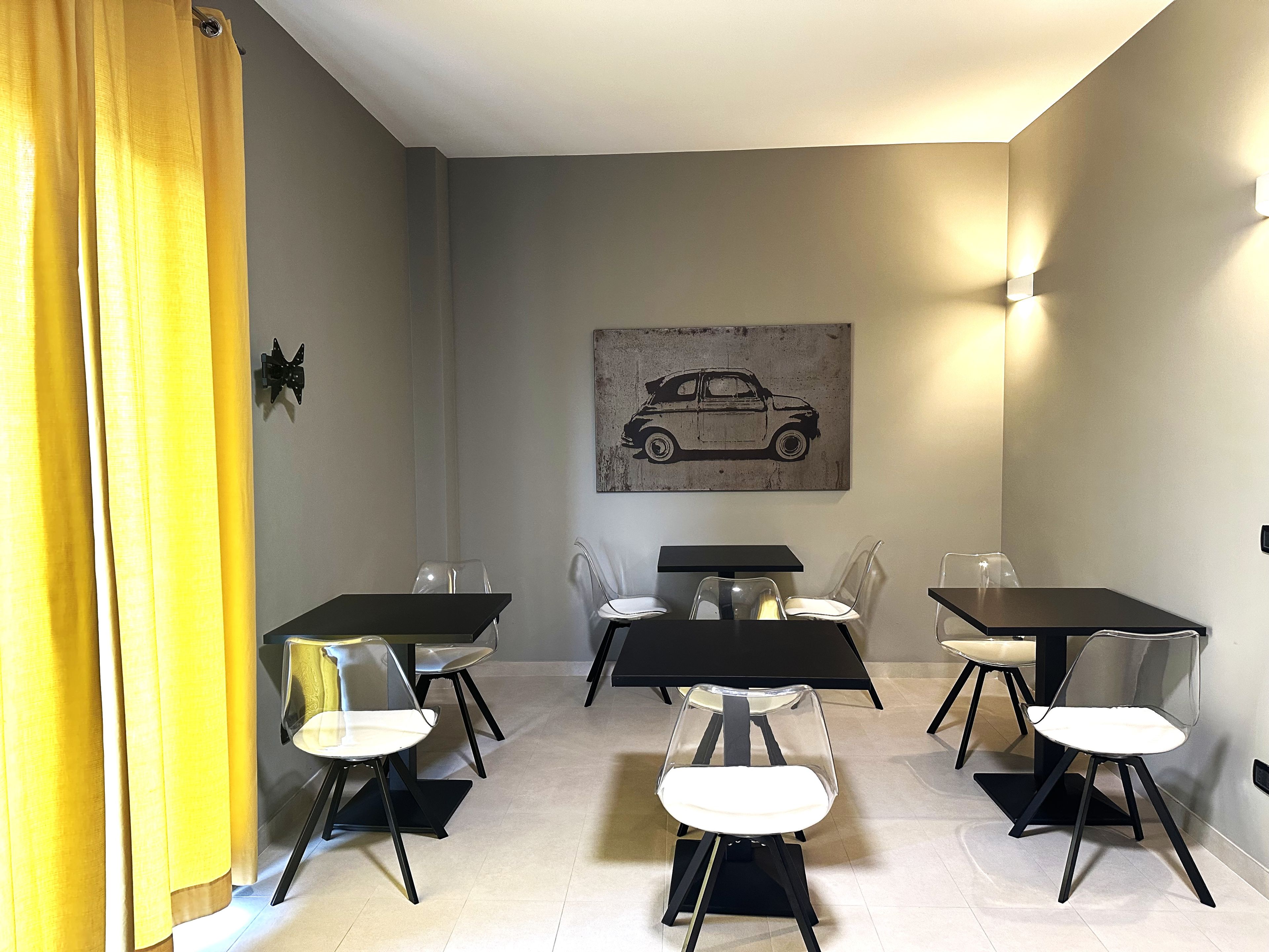 Un comedor con cuatro mesas y sillas, una cortina amarilla e ilustración de un coche colgada en la pared.