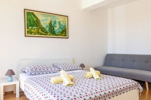 Un dormitori amb un llit, un sofà gris, un quadre d'un paisatge de muntanya i una tauleta de nit blanca amb una làmpada.