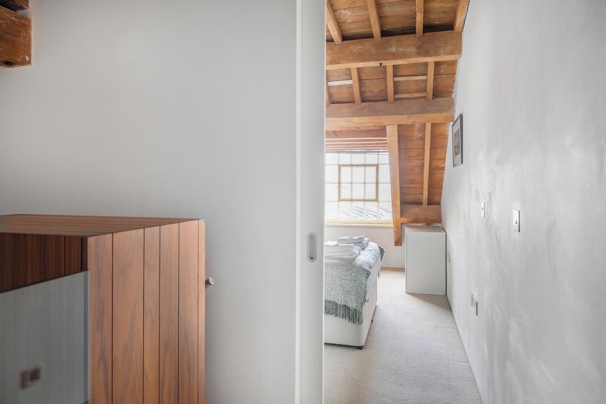 Ein schmaler Gang führt zu einem Schlafzimmer mit Holzbalkendecke und einem Fenster.