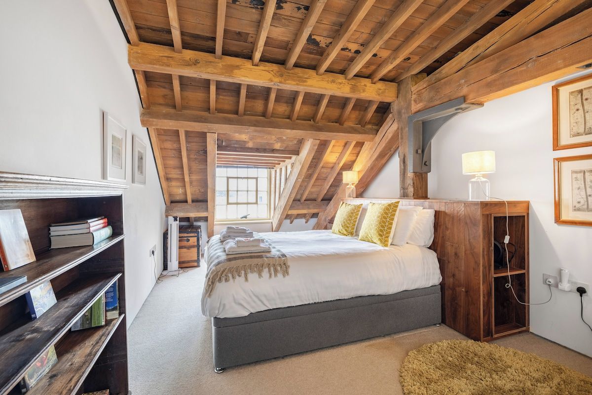 A bedroom with a wooden ceiling, a bed with pillows, a bedside table with a lamp, and a bookshelf.