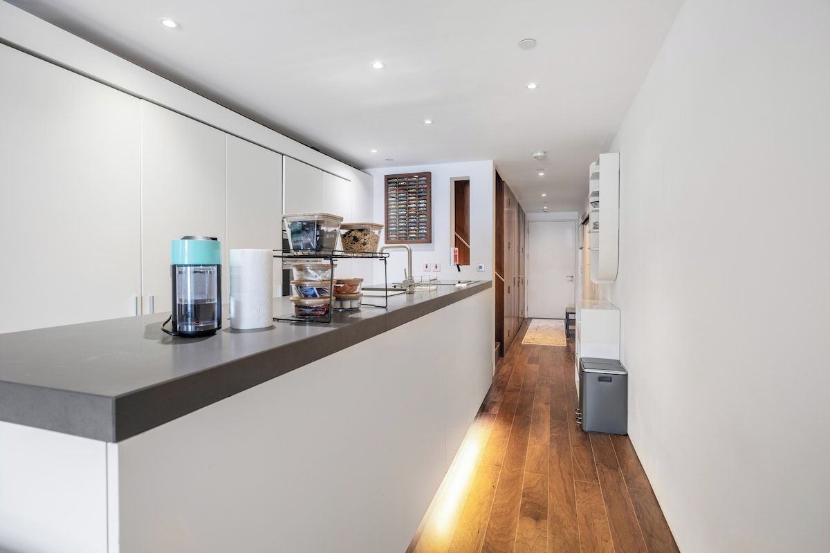A modern kitchen with a long countertop, a coffee machine, and a trash bin.