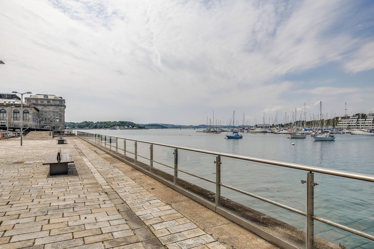 A waterfront promenade with a stone walkway, metal railing, and a row of boats.