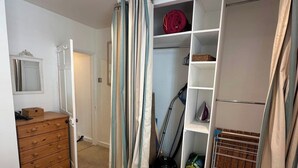 A bedroom with a bed, a wooden dresser, a closet with shelves, and a vacuum cleaner. - Dean Cottage - A Beautiful C17 Fisherman's Cottage (St Ives)