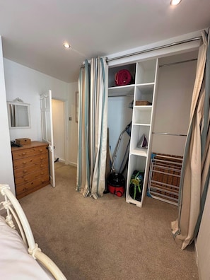 Un dormitorio con cama, cómoda de madera, armario con estantes y un aspirador.