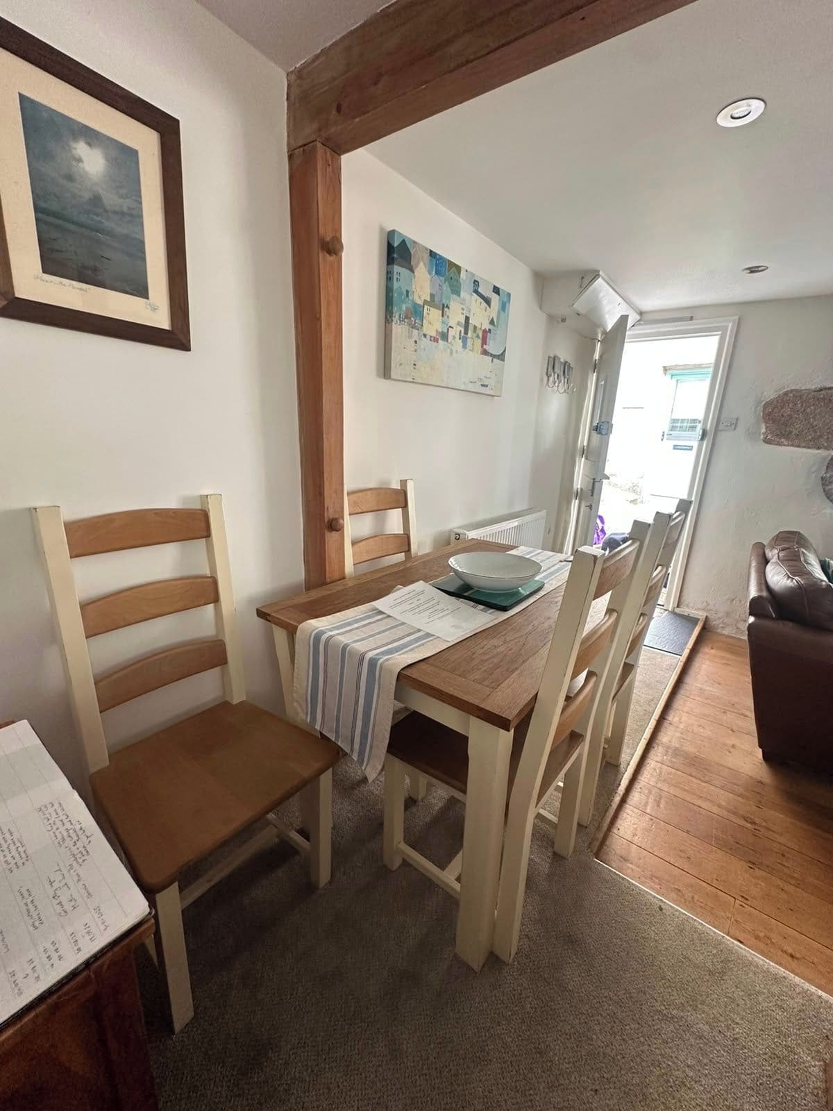 A dining area with a wooden table, chairs, and a bowl. There is a painting on the wall and a window with a view.