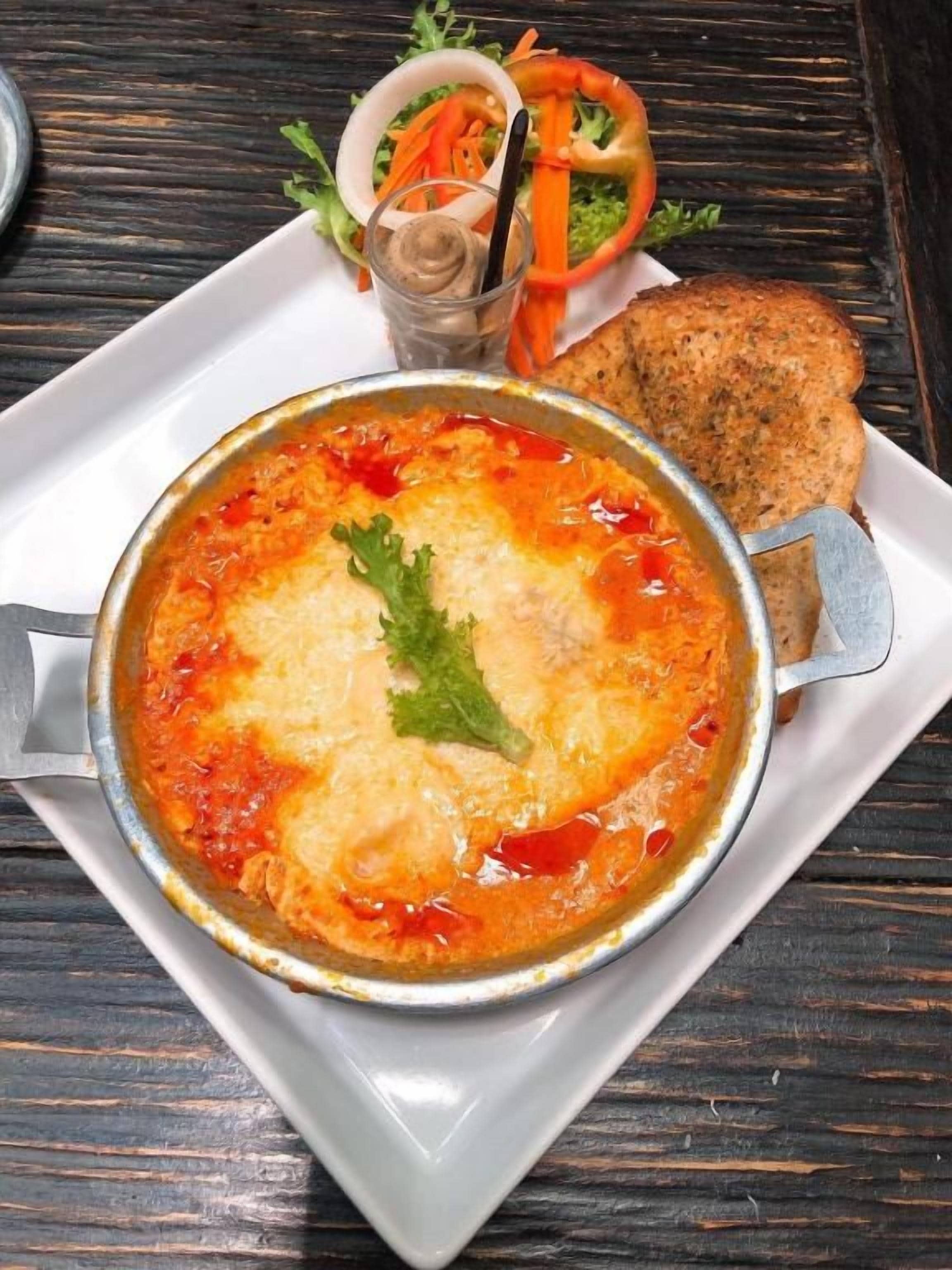 Eine Schüssel Suppe mit Garnitur auf einem weißen Teller, dazu eine Brotscheibe und ein kleiner Salat auf einem Holztisch.