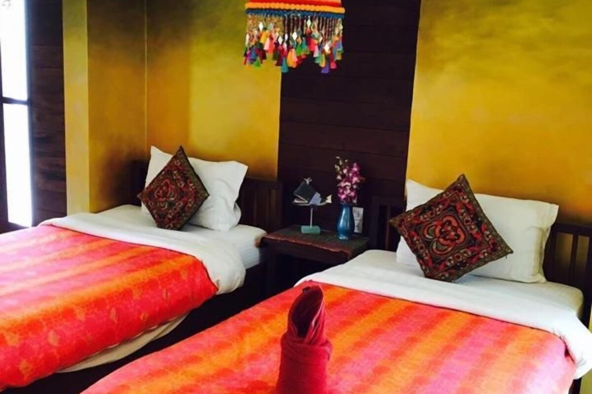 A hotel room with two beds, each with a red and orange striped bedspread, a hanging colorful lamp, and a small table with a vase of flowers.