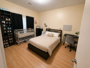 Ein Schlafzimmer mit einem Bett, einem Schreibtisch, einem Stuhl, einem Reisebett und einem Schuhregal.