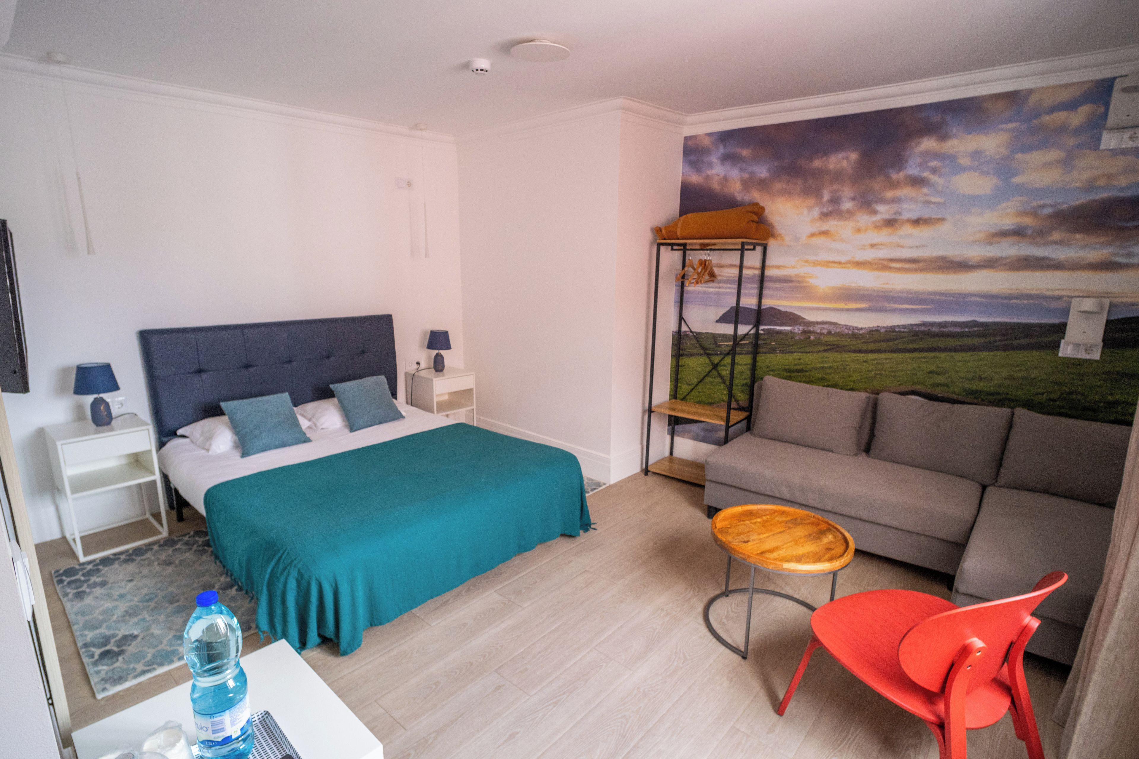 Un dormitorio con una cama, un sofá, una silla roja, una mesita y una pared con un cuadro de paisaje.