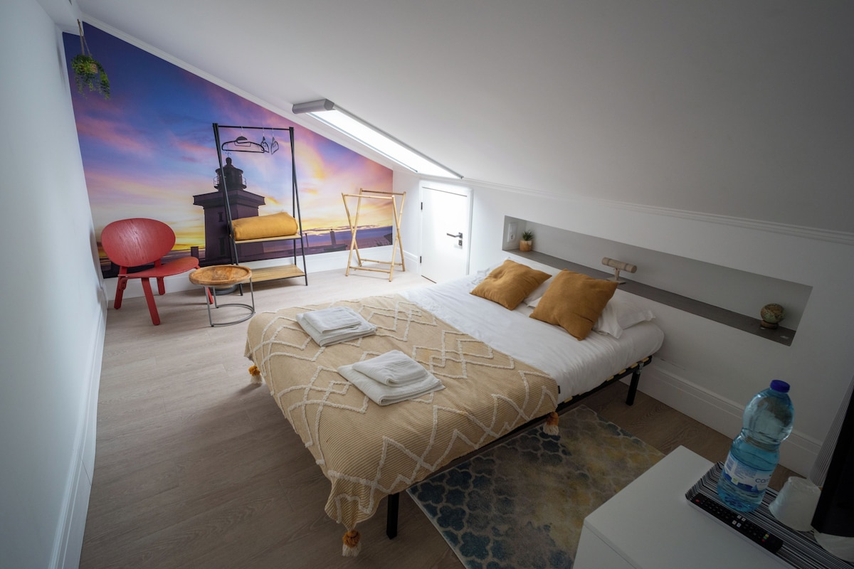 A bedroom with a bed, a chair, a hanging plant, a painting of a lighthouse, and a water bottle.