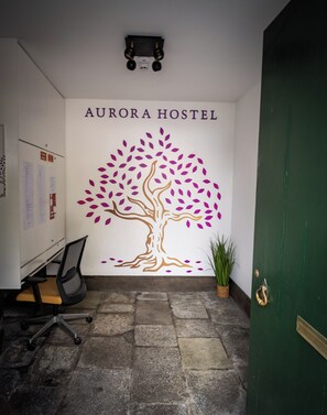 A wall with 'AURORA HOSTEL' text and a tree mural. There's a desk with a chair, a potted plant, and a green door.