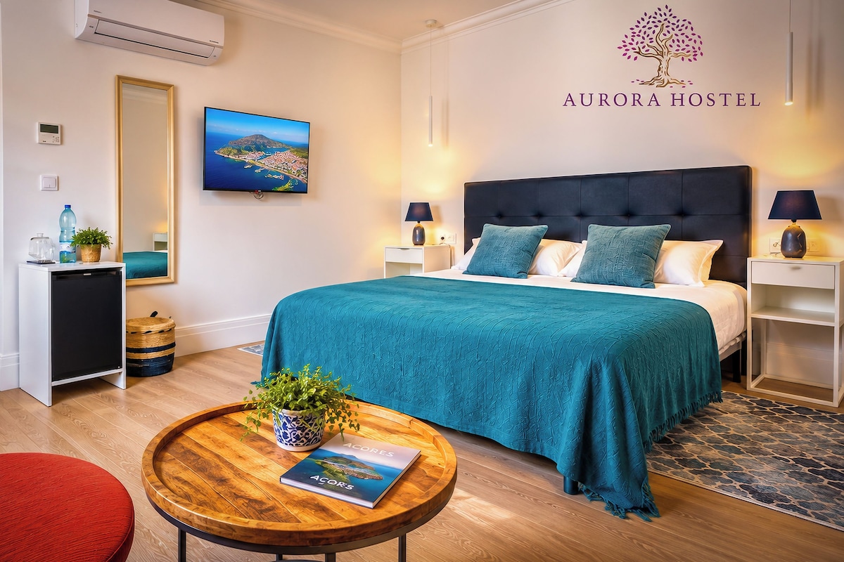 A hotel room with a bed, bedside tables, a round wooden table with a book, a potted plant, and a framed picture on the wall.
