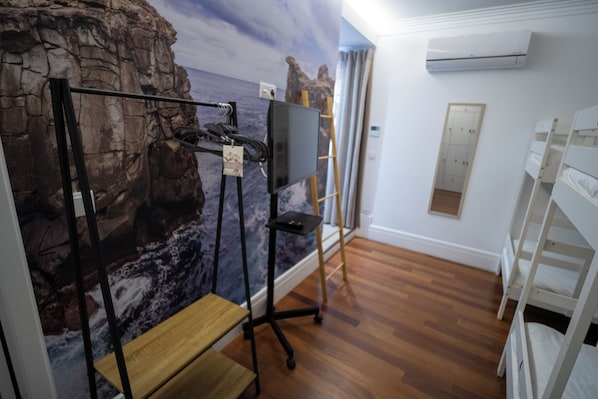 A room with a wooden floor, a desk with a chair, a mounted TV, and a wall with a rocky cliff mural.