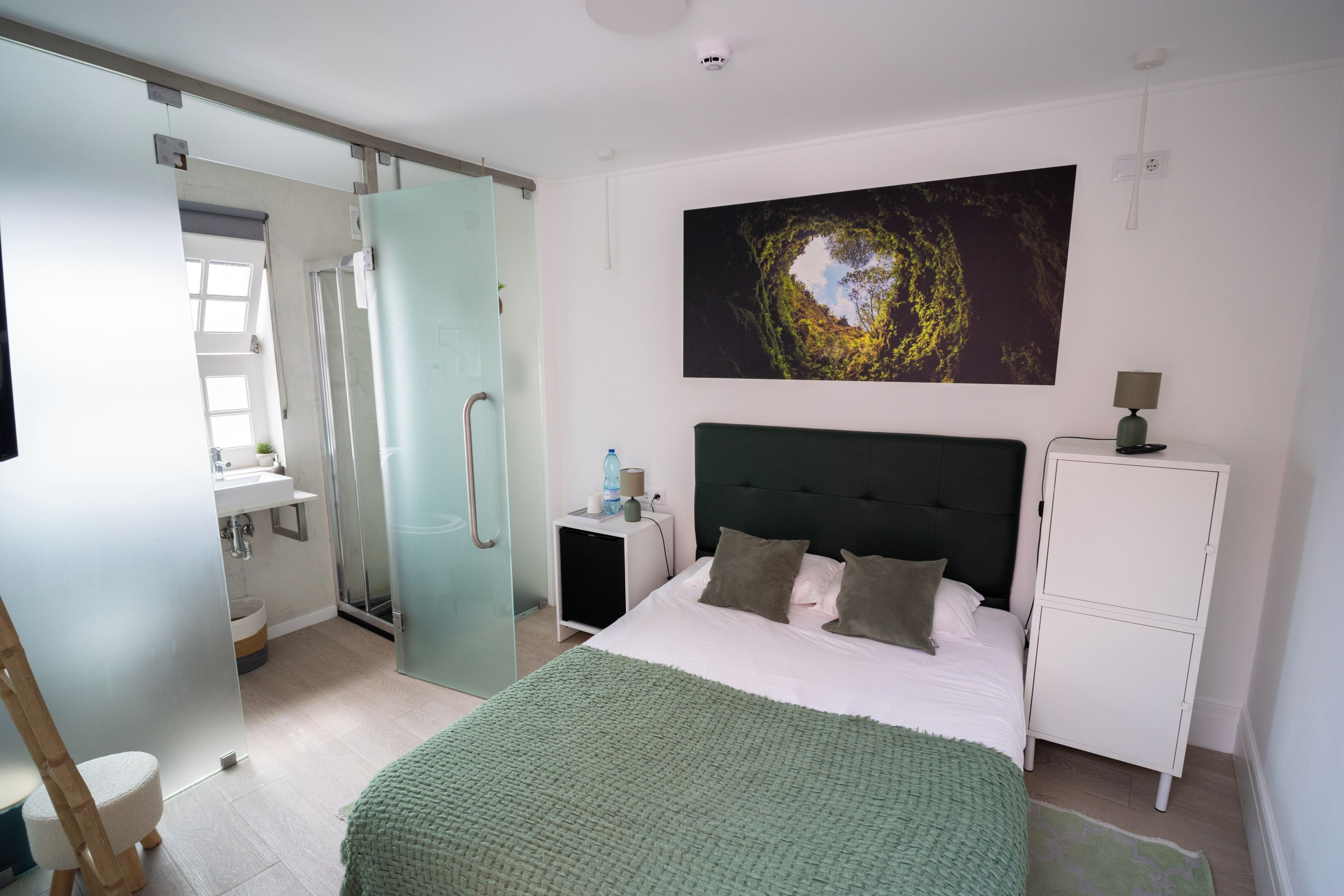 A bedroom with a bed, a nightstand, a dresser, a bathroom with a shower, and a large window.