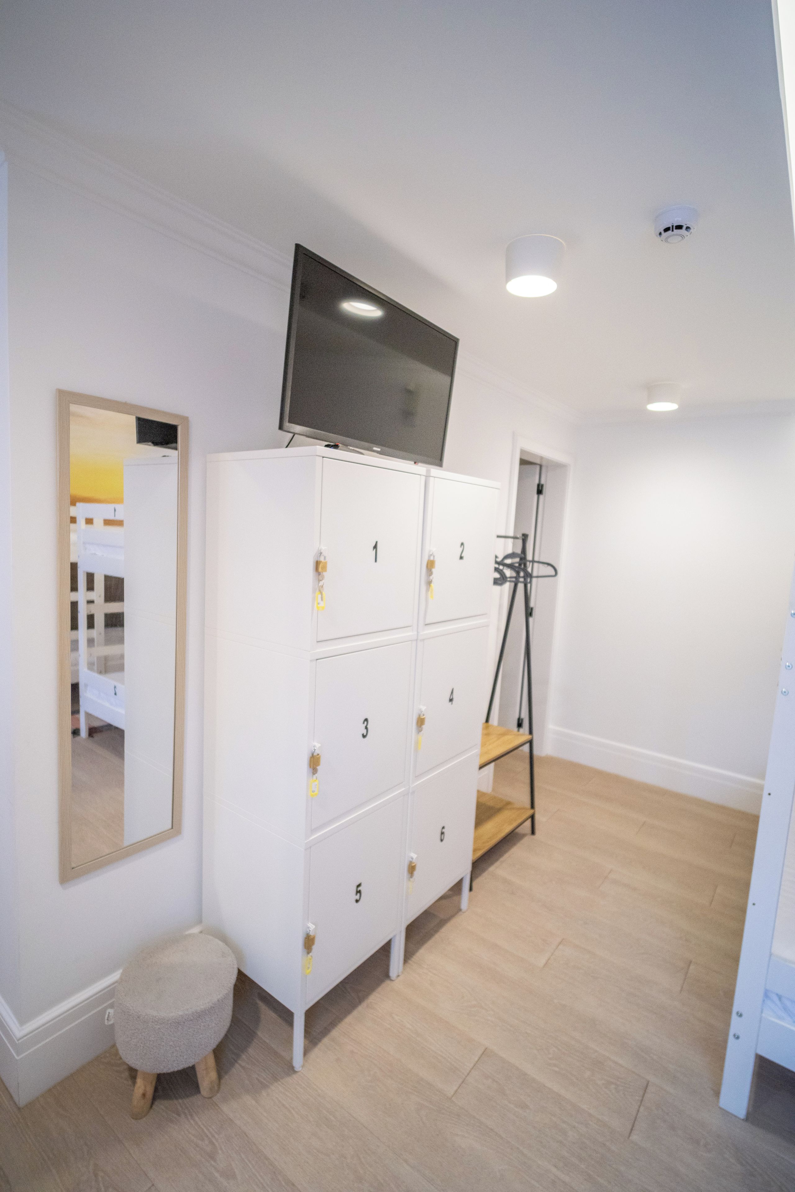 Una habitación con un armario blanco, un televisor sobre él, un taburete pequeño y un espejo en la pared.