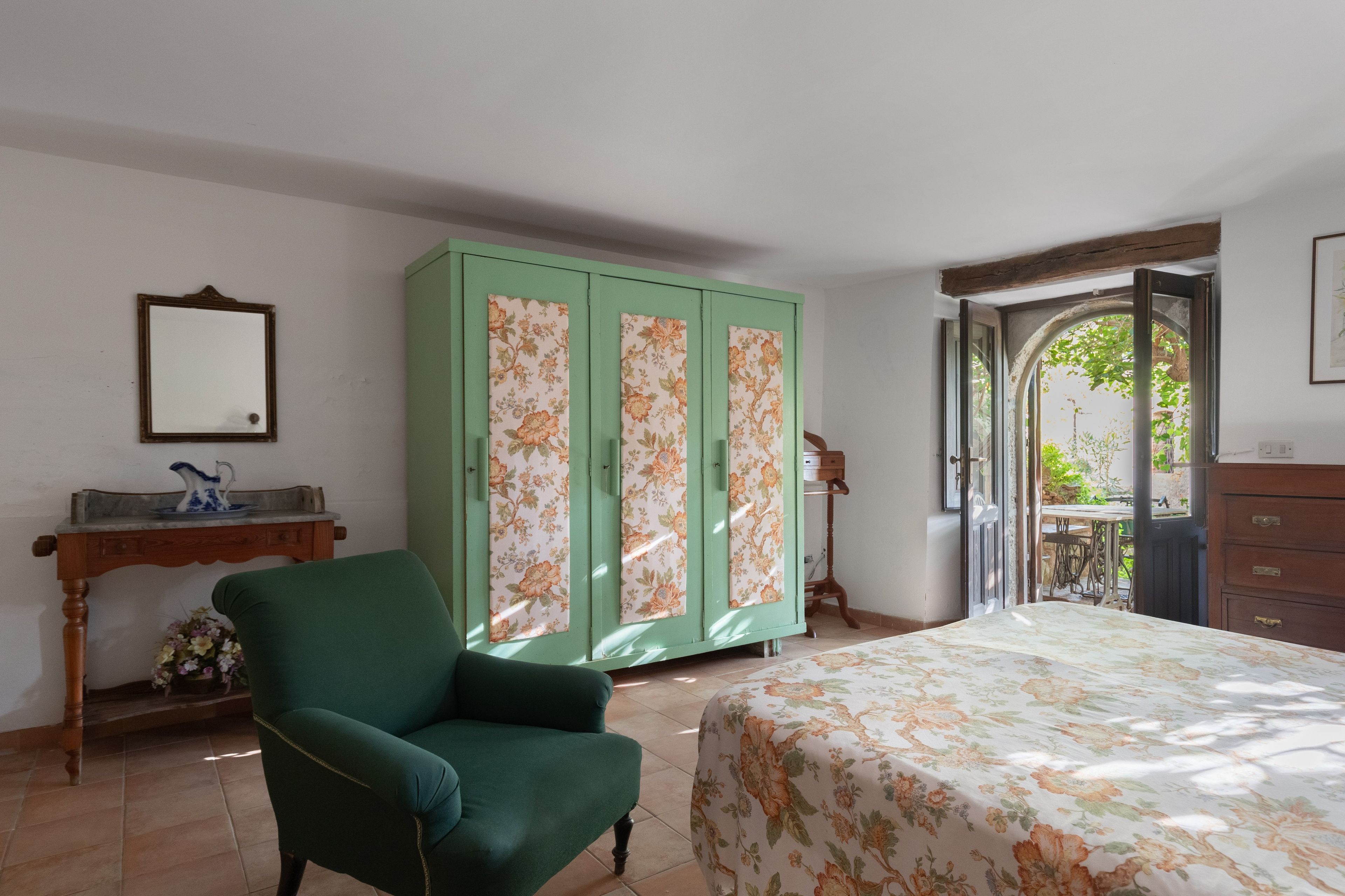 A room with a bed, a green armchair, a wooden dresser, a mirror, and a green wardrobe with floral panels.