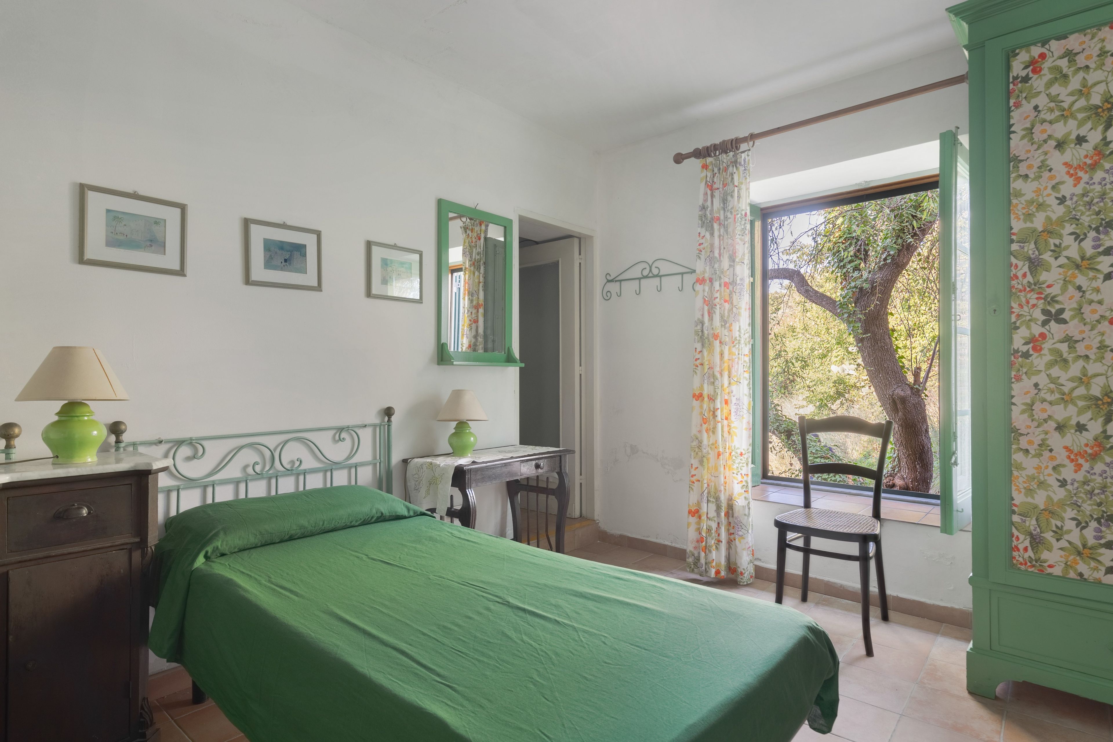 A bedroom with a bed, a chair, a desk, a lamp, and a window with a view of trees.