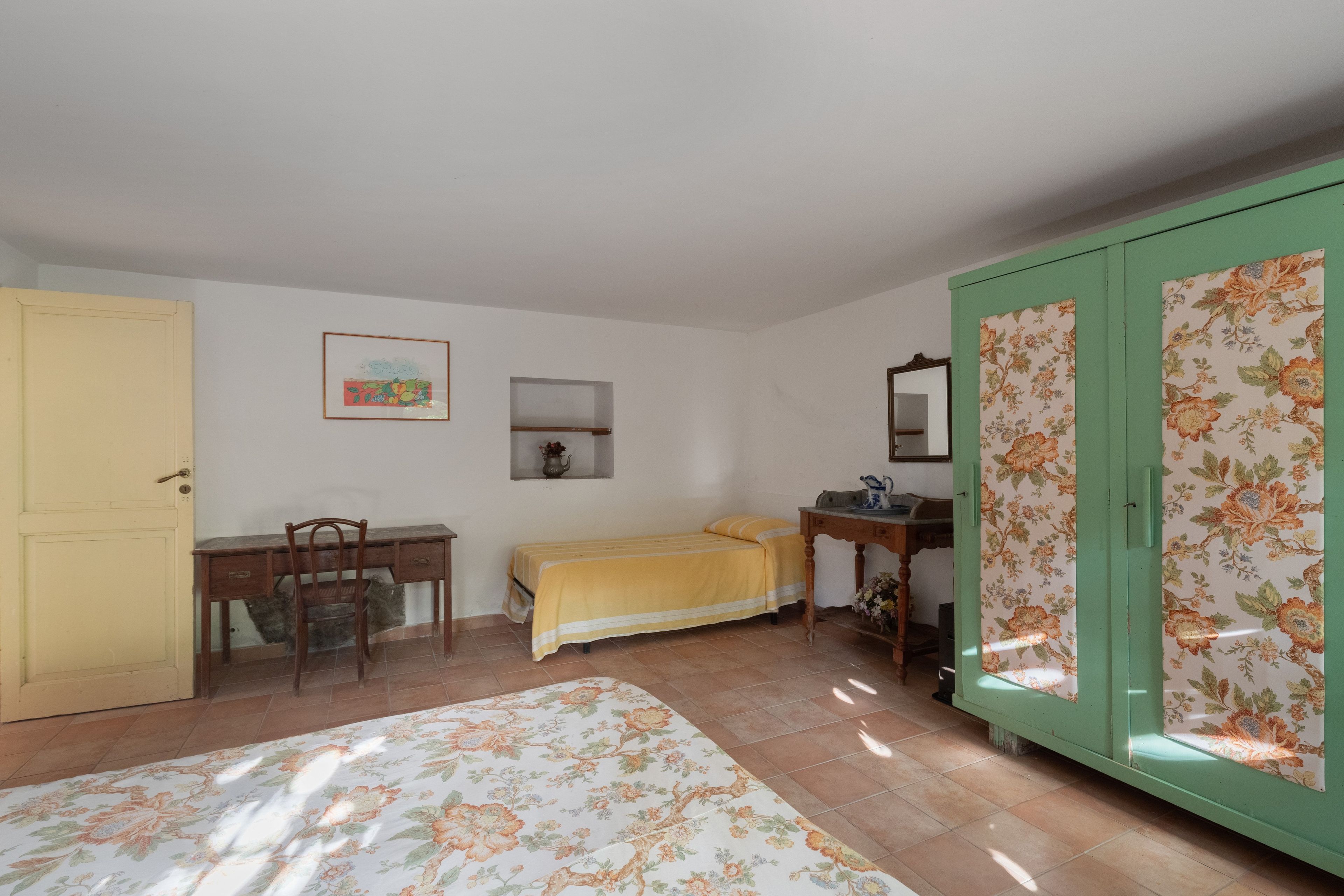 A room with a bed, a desk, a chair, a wardrobe, and a small shelf with a vase.