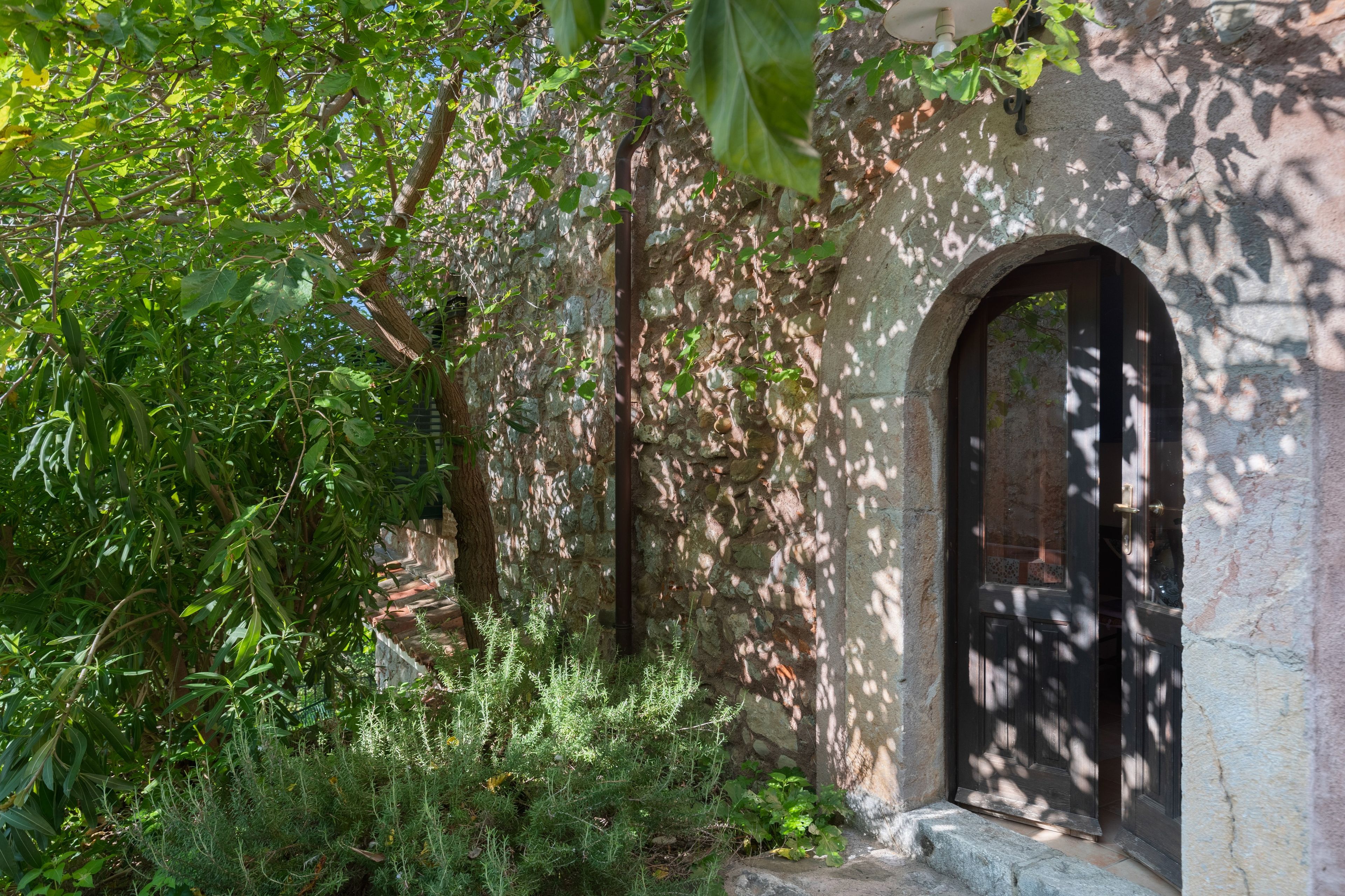 A stone building with an arched doorway, surrounded by dense foliage and a small garden area.