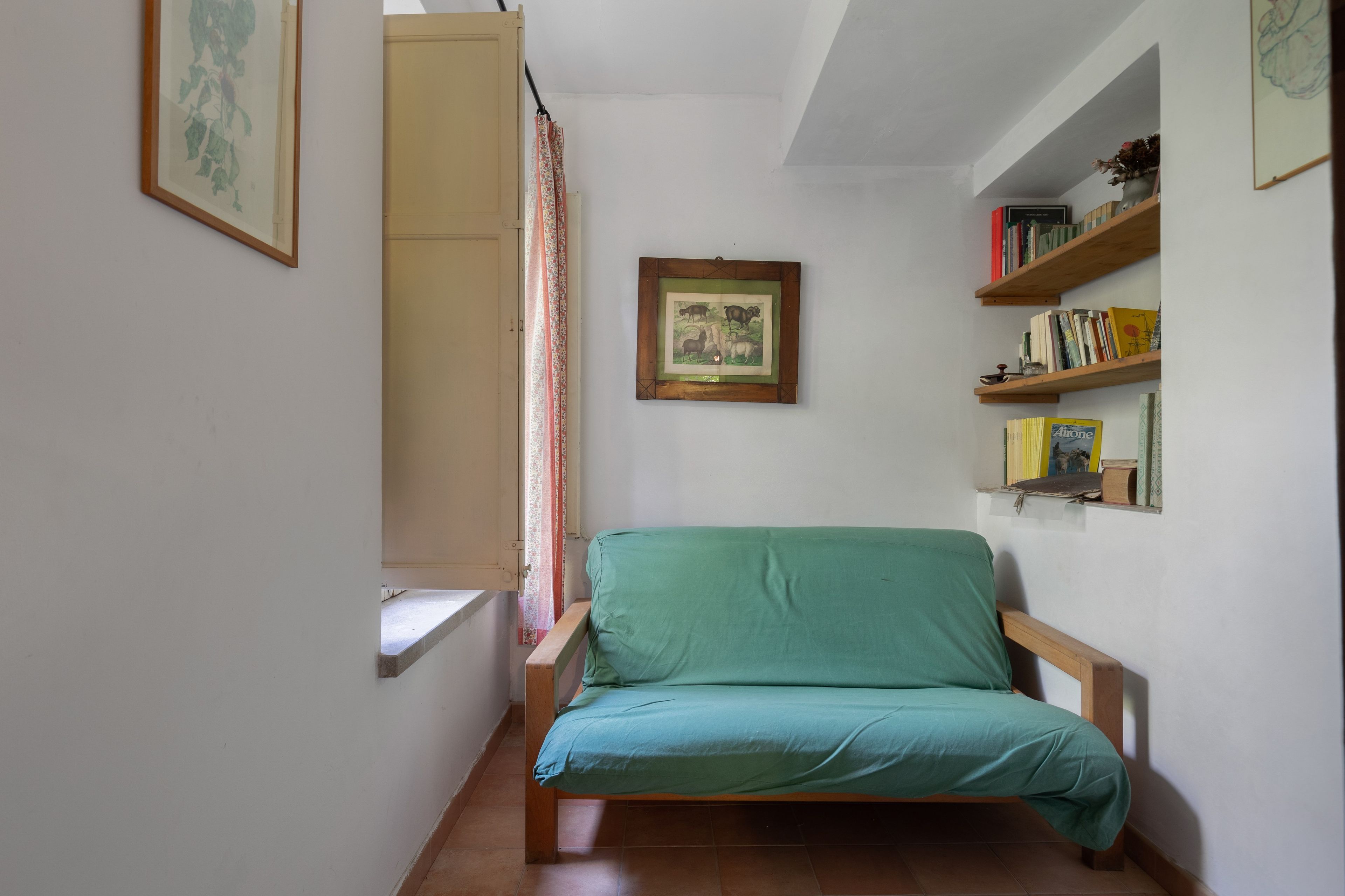A small, cozy room with a single bed, a window with curtains, a wall-mounted picture, and a bookshelf with books and decorative items.