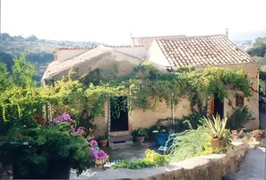 Un bâtiment rustique, doté d’un toit en tuiles, entouré d’une végétation luxuriante et de plantes en fleurs.