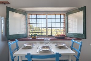 Mesa de jantar com cadeiras azuis, janela com vista para o verde e uma tigela de frutas no parapeito.