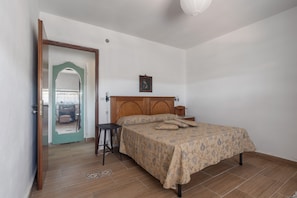Quarto com cama de madeira, mesa de cabeceira pequena, criadeira, um quadro na parede e porta que dá para outro cômodo.