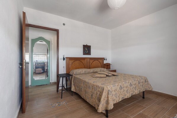 A bedroom with a wooden bed, a small bedside table, a nightstand, a wall-mounted picture, and a door leading to another room.