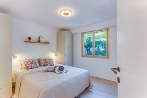 Una camera da letto con un letto, un comodino, una mensola a parete con oggetti decorativi, una finestra con vista sul verde e una porta con maniglia moderna.