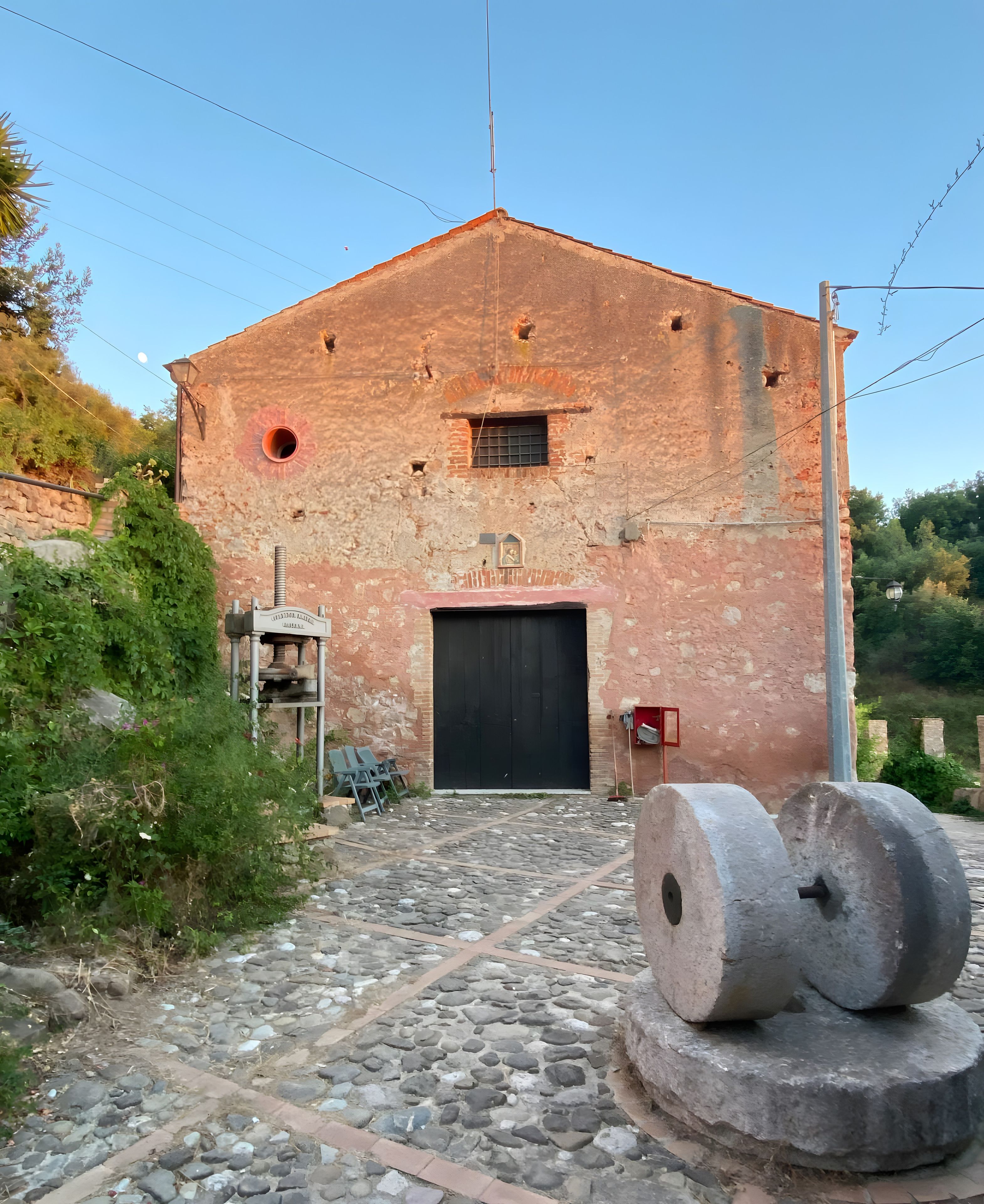 Un vieux bâtiment avec une grande porte noire, une petite fenêtre et une cloche. Devant la porte, on trouve deux grandes roues en pierre.