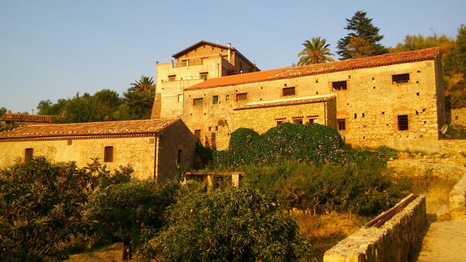 A stone building with multiple windows and a tiled roof, surrounded by greenery and a stone wall.