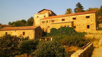 A stone building with multiple windows and a tiled roof, surrounded by greenery and a stone wall.